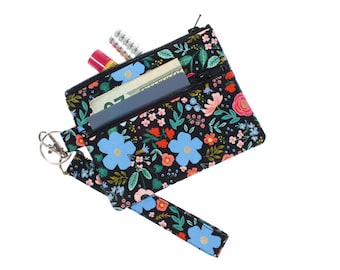 May include: A black zippered pouch with a floral print featuring blue, pink, red, and green flowers. The pouch has a key ring attached and is partially open, revealing a blue card, cash, and a red lip balm.