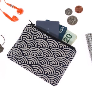May include: A small, black and white patterned zippered pouch with a blue and white credit card and cash inside. The pouch is lying on a white surface with a pair of orange earbuds, two keys, and a few coins.