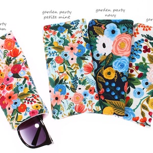 May include: Four floral patterned eyeglass cases in cream, mint, navy, and cream. The cases are lined up horizontally with the cream case on the right and the mint case on the left. A pair of sunglasses is partially visible in the cream case.