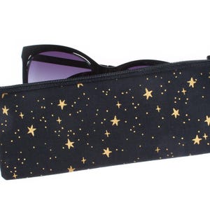 May include: Black eyeglass case with a gold star pattern and a black zipper. The case holds a pair of sunglasses with black frames and purple lenses. A silver ring is attached to the side of the case.
