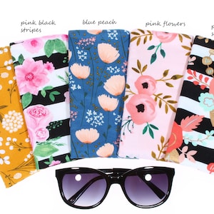 May include: Six different fabric eyeglass cases with floral patterns. The cases are arranged in a row with a pair of black sunglasses in the middle. The cases are labeled with the following names: mustard yellow, pink black stripes, blue peach, pink flowers, peach black stripes.
