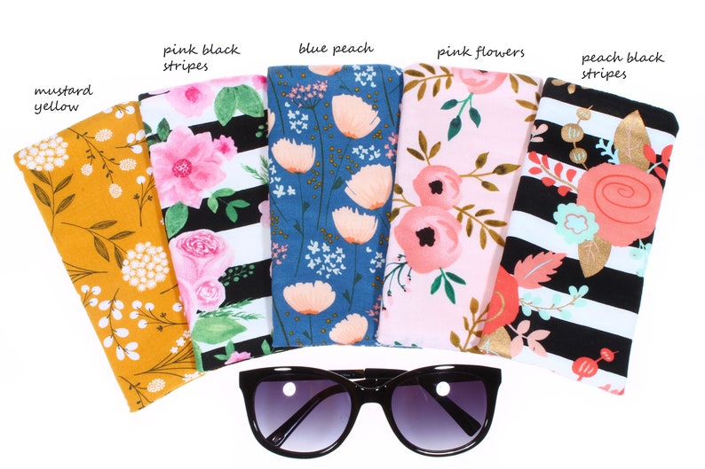 May include: Six different fabric eyeglass cases with floral patterns. The cases are arranged in a row with a pair of black sunglasses in the middle. The cases are labeled with the following names: mustard yellow, pink black stripes, blue peach, pink flowers, peach black stripes.