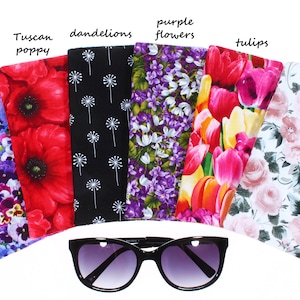 May include: Six different floral patterned eyeglass cases, each with a different flower design. The cases are arranged in a semi-circle with a pair of black sunglasses in the center. The cases are labeled with the names of the flowers: pansies, Tuscan poppy, dandelions, purple flowers, tulips, and roses.