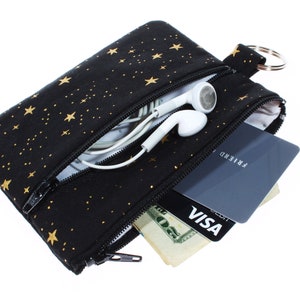 May include: A black zippered pouch with a gold star pattern. The pouch is open and contains a credit card, cash, and a pair of white earbuds.