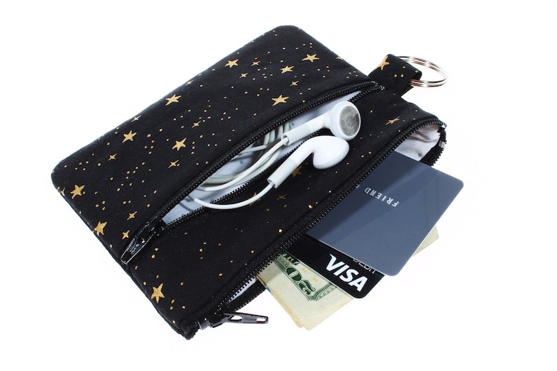 May include: A black zippered pouch with a gold star pattern. The pouch is open and contains a credit card, cash, and a pair of white earbuds.