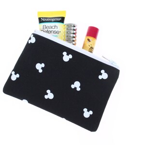 May include: Black zippered pouch with a white Mickey Mouse pattern. The pouch is partially open and contains a yellow sunscreen bottle with the text "Neutrogena Beach Defense", a silver tube of lip balm, and a red tube of lip balm.