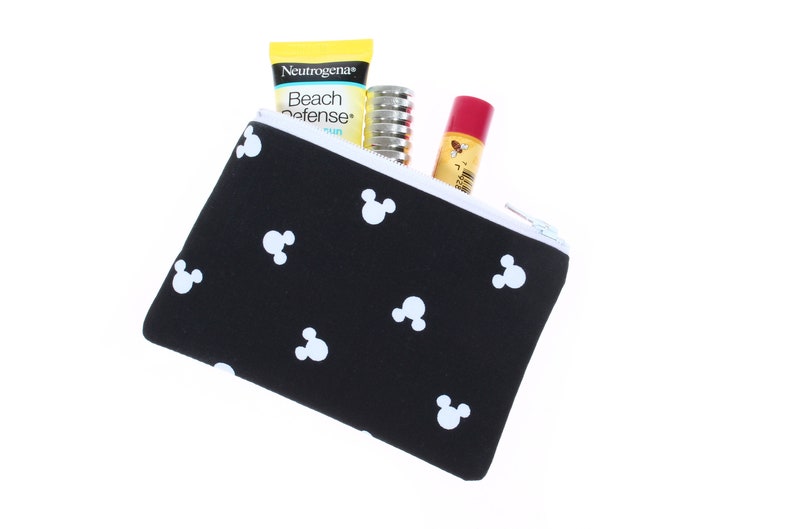May include: Black zippered pouch with a white Mickey Mouse pattern. The pouch is partially open and contains a yellow sunscreen bottle with the text "Neutrogena Beach Defense", a silver tube of lip balm, and a red tube of lip balm.