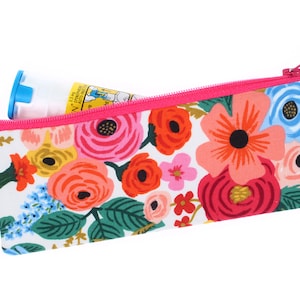 May include: A pink zippered pouch with a floral print featuring pink, red, yellow, and blue flowers on a white background. The pouch has a silver key ring attached and a small blue and white tube is partially inserted into the pouch.