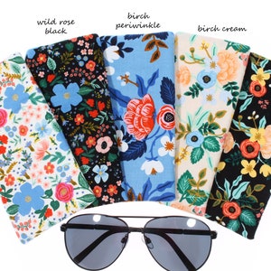 May include: Five different floral patterned eyeglass cases, each with a different color background: white, black, blue, cream, and black. The cases are arranged in a semi-circle with a pair of black sunglasses in the center.