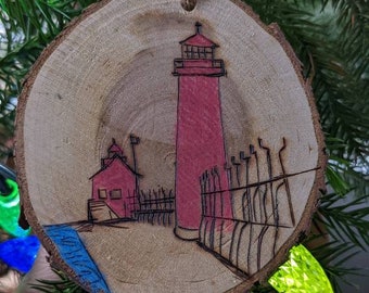 May include: A wooden ornament with a hand-painted image of a red lighthouse on a blue body of water. The ornament is hanging from a string on a green Christmas tree.
