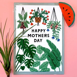 Puede incluir: Una tarjeta de felicitación con borde verde y el texto "HAPPY MOTHERS DAY". La tarjeta presenta ilustraciones de plantas colgantes, hojas tropicales y una rodaja de sandía. La paleta de colores incluye verde, blanco, rojo y rosa.