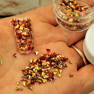 May include: A hand holding a small clear plastic container and a white lid. The container holds small, shiny, red and gold triangle-shaped glitter. Some glitter is scattered in the hand and in a small plastic container.