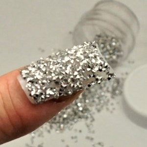 May include: A fingernail is covered in silver glitter, with a small jar of loose silver glitter in the background.