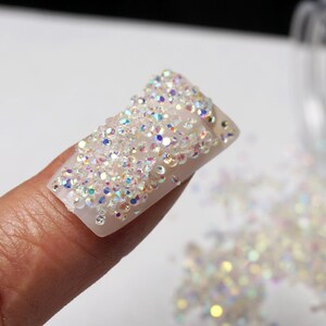 May include: A close-up of a fingernail covered in iridescent rhinestones. The nail is rectangular and the rhinestones are scattered across the surface. A clear container is out of focus in the background, with more rhinestones scattered around.