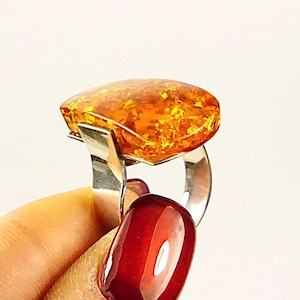 May include: A ring featuring a large, polished, translucent amber stone with golden and orange hues. The stone is set in a smooth, wide silver band that curves upward to support the base of the amber, creating a distinct profile view of the jewelry.