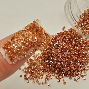 May include: A clear plastic jar filled with small, iridescent copper-colored rhinestones. The rhinestones are scattered on a white surface.
