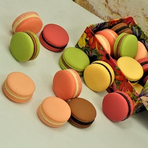 May include: A selection of colourful macarons in shades of pink, orange, yellow, green, and brown. Some macarons are scattered on a white surface, others are in a floral patterned container. The macarons have a cream filling.