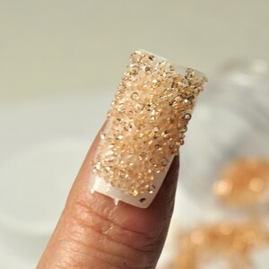 May include: A close-up of a finger with a clear nail with a layer of small, gold-colored beads on top.