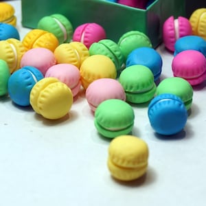 May include: A collection of colourful macarons in various shades of blue, green, yellow, pink, and white. The macarons are arranged on a white surface.