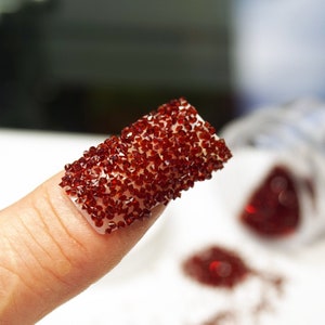 May include: A fingernail with a red glitter nail art design. The glitter is applied in a thick layer and covers the entire nail. The glitter is a mix of small and large pieces.