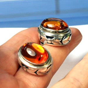 May include: Two oval-shaped amber rings with a silver band. The amber stones have a warm, orange-brown hue. The silver bands feature a decorative leaf design. The rings are displayed on a hand.