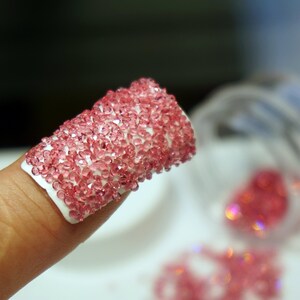 May include: A fingernail with a pink rhinestone design. The rhinestones are small and closely packed together, creating a sparkling effect. The rhinestones are also scattered on a white surface.