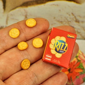 May include: A miniature box of Rizz crackers, a popular brand of snack crackers, with a red background and white text. The box is labeled "Rizz" and "Ritz Crackers". There are six miniature crackers in the image, all of which are round and yellow.