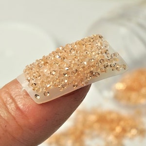 May include: A close-up of a finger wearing a fake nail with a design of small, sparkly, light brown rhinestones.