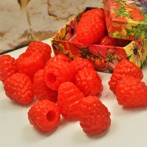 May include: A box filled with bright red, realistic-looking raspberry-shaped candies. The box is decorated with a floral pattern. The candies are scattered around the box on a white surface.