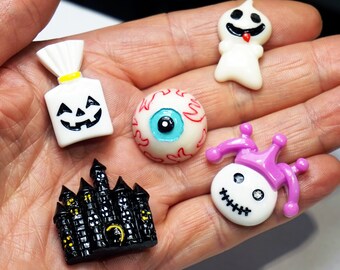 May include: Five small resin cabochons featuring Halloween themes: a white ghost, a white jack-o'-lantern treat bag, a realistic eyeball with red veins, a black haunted castle with yellow windows, and a white jester head with a purple hat.