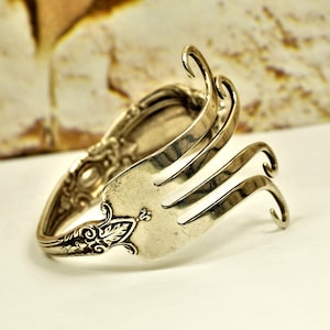 May include: Silver-toned fork bracelet. The bracelet is crafted from a repurposed fork, with the tines bent upwards to create a unique design. The handle features ornate detailing, adding a vintage aesthetic.