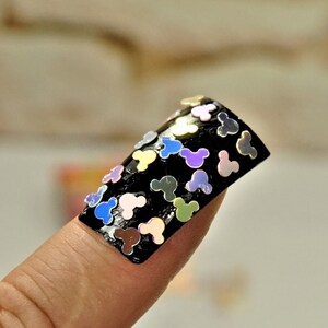 May include: A black nail with iridescent Mickey Mouse head-shaped sequins. The sequins are in various colors, including blue, pink, green, and yellow. The nail is on a finger.