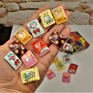 May include: A selection of small, colourful food-themed charms, including packets of cheese, strawberries, and cookies. The charms feature detailed graphics and text, with some displaying the words "COOKIES" and "CHEESE".