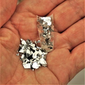 May include: A hand holding a small pile of silver glitter and a sheet of silver glitter on a clear plastic backing.