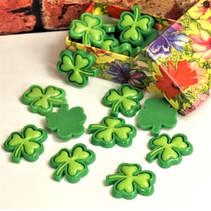 May include: A collection of small, bright green shamrock-shaped buttons scattered on a white surface, with several more contained inside a small, rectangular box decorated with a colorful floral pattern of red, yellow, and purple flowers.