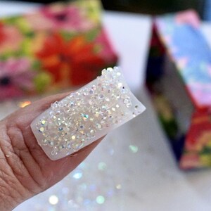 May include: A white acrylic nail covered in iridescent rhinestones. The nail is held up, showcasing the sparkling crystals. The background includes blurred floral-patterned boxes, suggesting a nail art or beauty product.