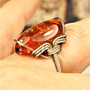 May include: A ring featuring a polished, oval-shaped amber gemstone with a deep orange hue. The stone is secured by a silver-toned metal wire wrap design that coils around the base and sides, creating a textured, industrial-style band structure.