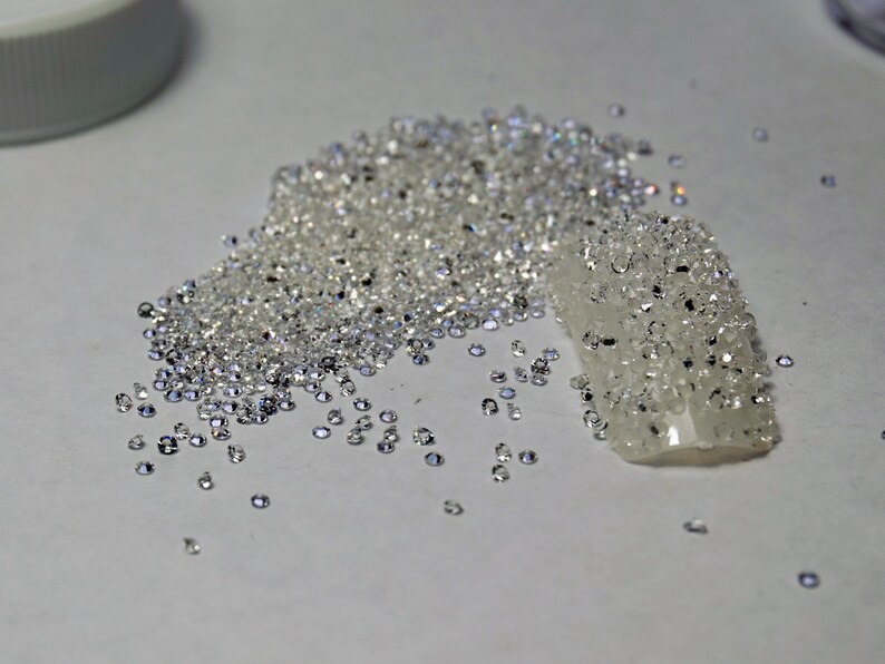 May include: A pile of small, clear, iridescent rhinestones, some of which are scattered on a white surface. A small, clear, plastic strip is covered in rhinestones.