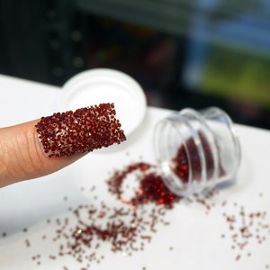 May include: A fingernail covered in small, dark red rhinestones. A clear plastic container of rhinestones is tipped over, spilling the gems onto a white surface. A white lid is also visible.