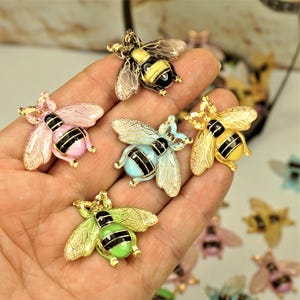 May include: Five bee-shaped charms in various colours including yellow, pink, light blue, and green. Each charm features gold-toned wings with a detailed filigree pattern, a black and gold striped body, and a small loop at the top for attachment.