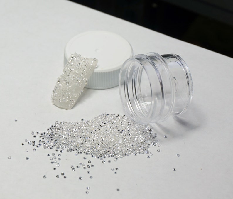 May include: A clear plastic jar with a lid, filled with small, white, iridescent beads. Some beads have spilled out of the jar and are scattered on a white surface.