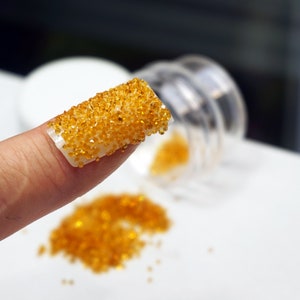 A close-up of a finger with a clear nail polish base and a layer of small, round, orange-yellow rhinestones.