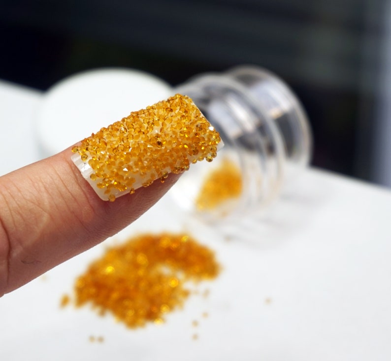A close-up of a finger with a clear nail polish base and a layer of small, round, orange-yellow rhinestones.