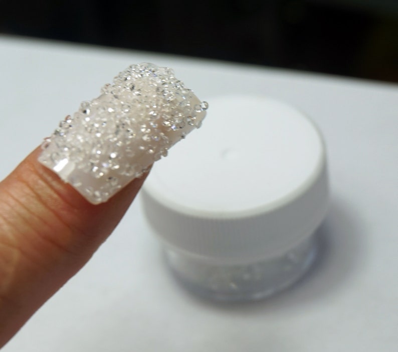 May include: A fingernail with a clear base and a layer of small, clear, iridescent glitter on top. The glitter is in a small, clear jar with a white lid.