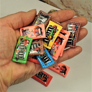 May include: A collection of miniature M&M's candy bar replicas in various colours, including pink, green, yellow, and blue. Each candy bar features the M&M's logo and character designs. The candies are small, fun-sized, and appear to be made of resin.