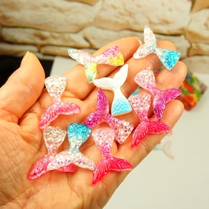 May include: A collection of small, decorative mermaid tail charms in various colours including pink, blue, yellow, and iridescent white. Each charm features a textured, glittery finish with a distinct fin shape, crafted from a smooth, hard material.
