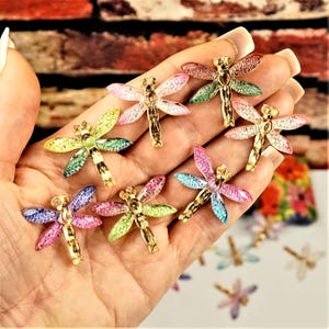 May include: A collection of small dragonfly-shaped charms featuring gold-toned metal bodies and translucent, glittery wings in various colours including pink, yellow, green, blue, and purple, held in a palm for scale and display.