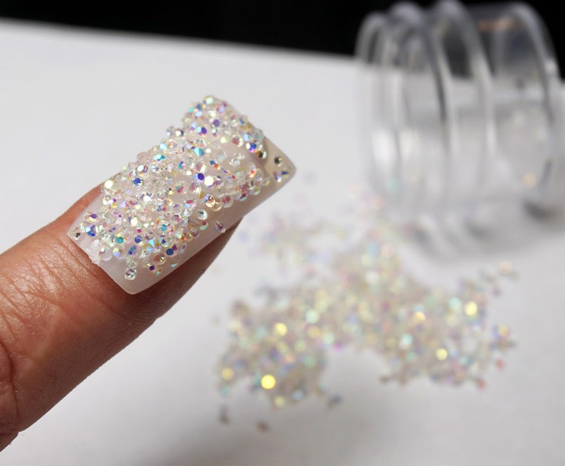 A close-up of a fingernail covered in iridescent clear rhinestones. The rhinestones are small and round, and they are arranged in a random pattern. The rhinestones are scattered around the fingernail, and they are also scattered on a white surface.