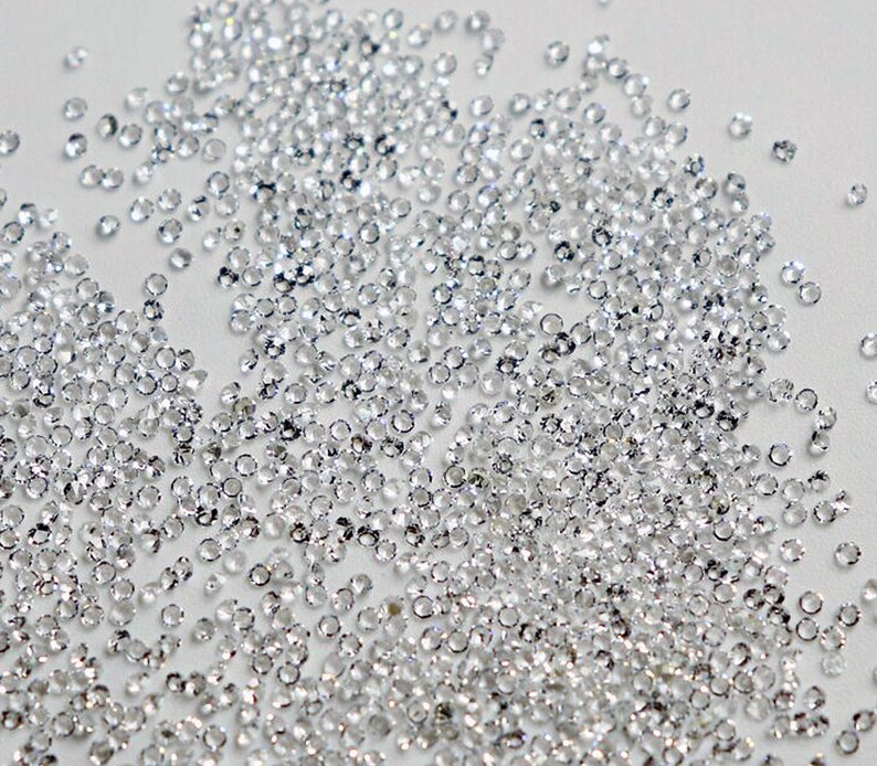 May include: A large quantity of small, clear, faceted rhinestones scattered on a white surface.