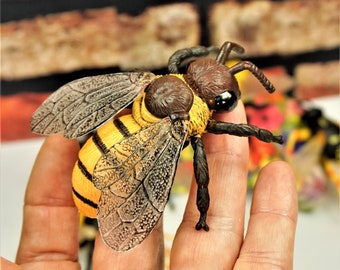 May include: A realistic-looking toy bee with brown and black legs, a yellow and black striped abdomen, and brown and black wings. The bee is sitting on a person's hand.